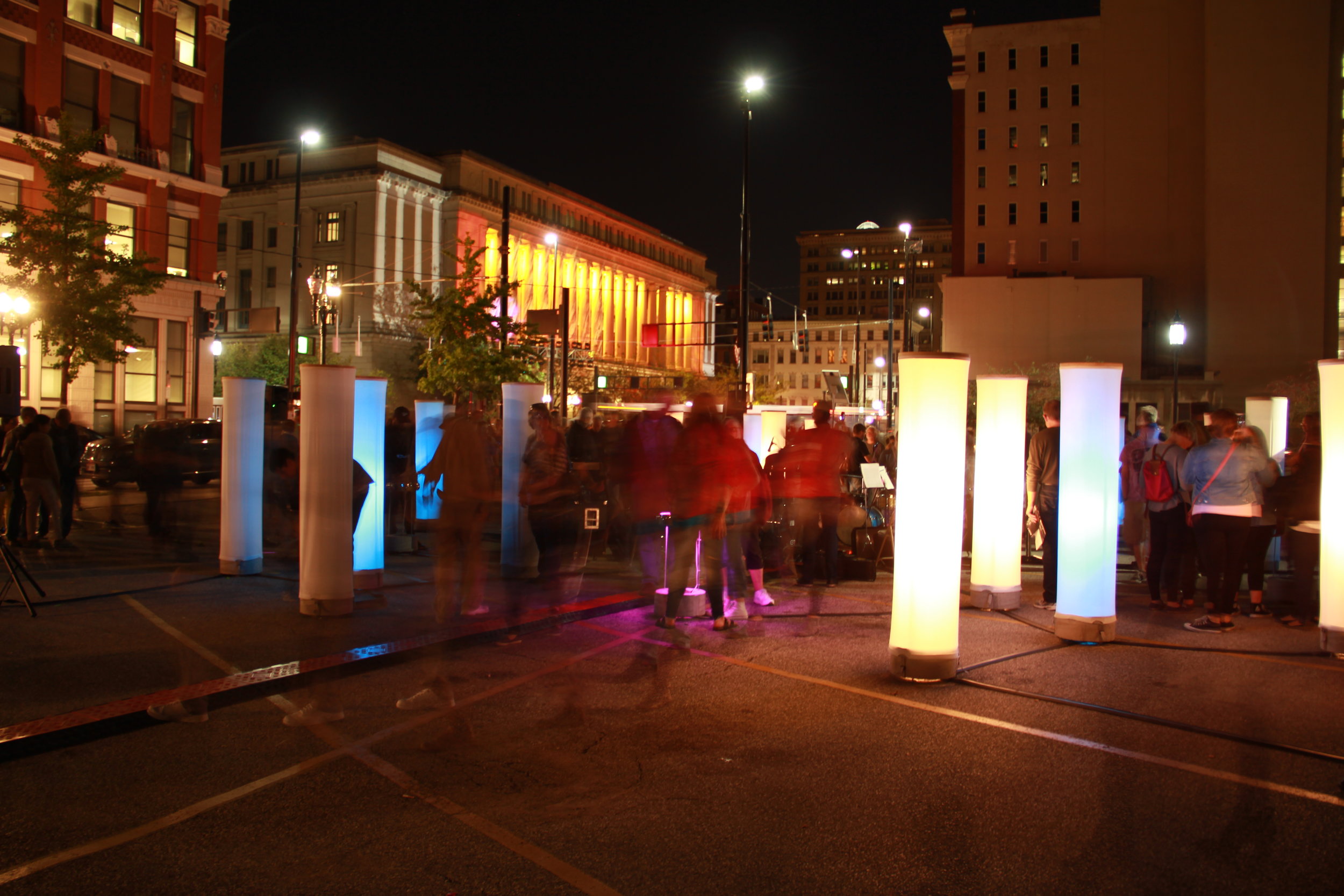 Ground-level night view of glowing fabric columns scattered across a downtown Cincinnati street with blurred figures moving through the installation