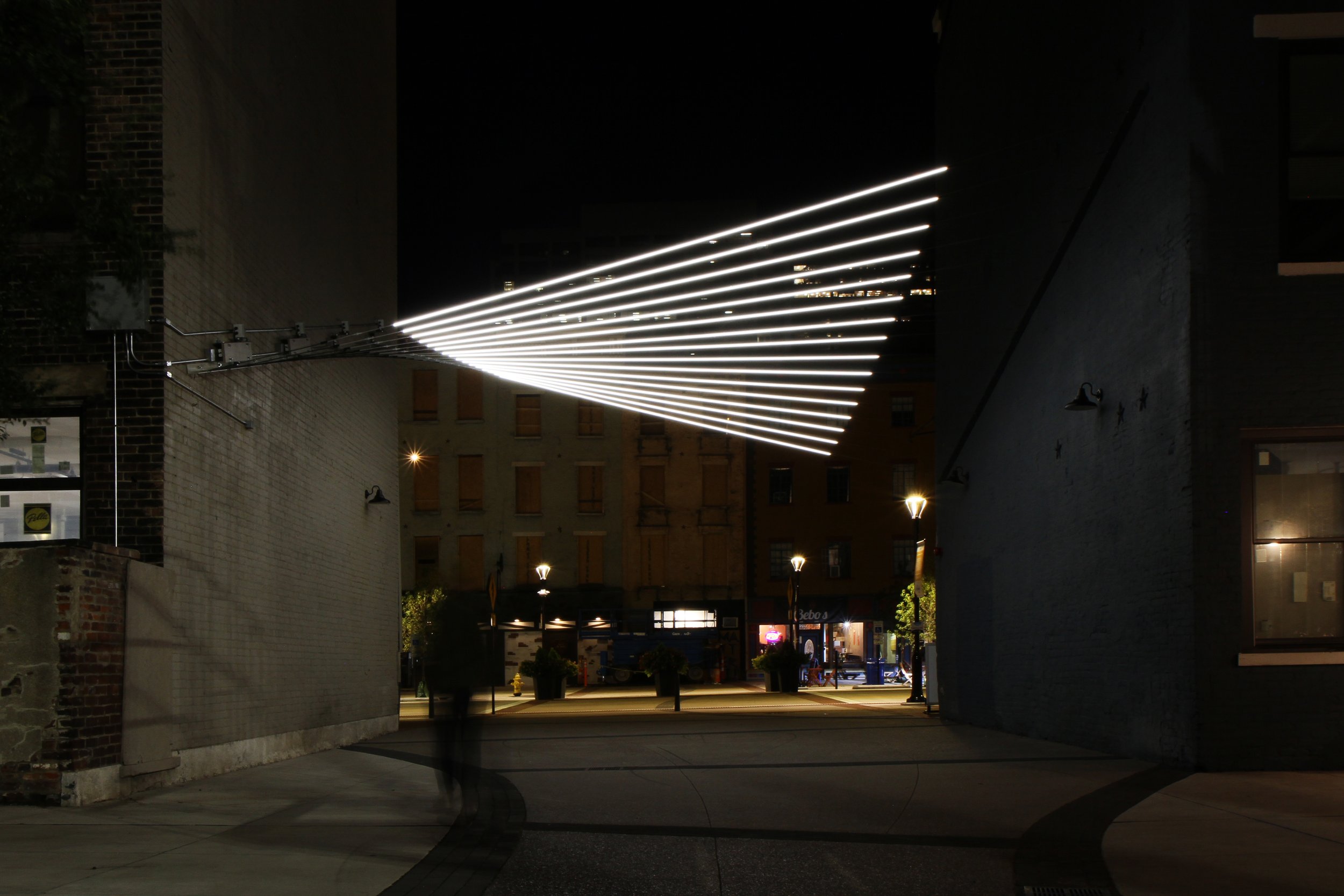 LED lines fanning from a narrow point on one building wall across the plaza gap, viewed from the street at night